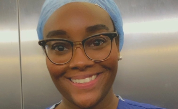 Dr Rochelle Pierre smiles at the camera wearing dark blue scrubs and a light blue hair cap. She has on glasses with rimmed lenses and a bright smile.